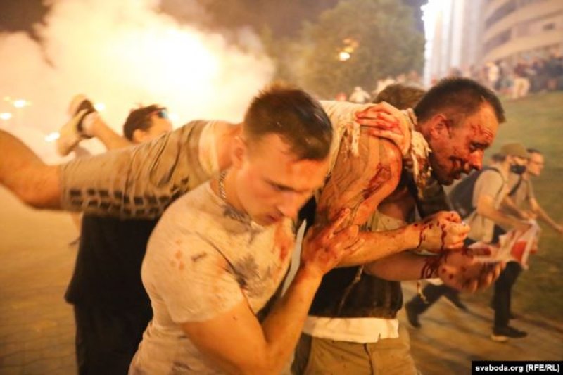 To menn som bærer en svært skadd mann med rennende blod overalt som viser Lukasjenko sin krigføring mot folket. Bilde er tatt på en protest mut Lukashenko og hans regime, som gikk fra fredelig til voldelige og farlig etter myndighetenes innblanding.